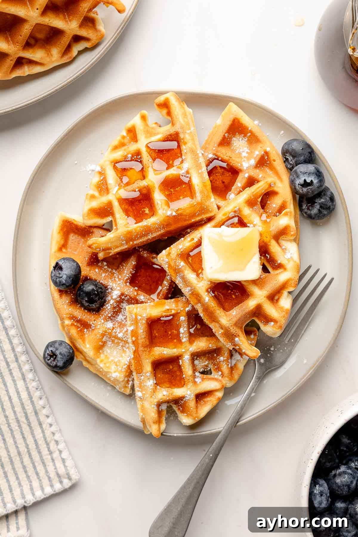 Gluten-free Belgian waffles on a gray plate topped with butter, maple syrup, and powdered sugar, served with fresh blueberries.