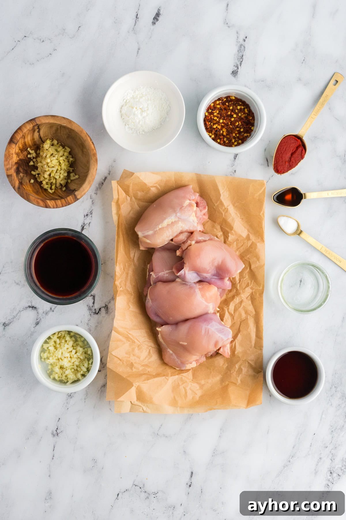 Sticky Air Fryer Chicken Thighs (Oven Method Included) 4 raw boneless skinless chicken thighs on parchment paper all on a marble background with all the ingredients for the marinade and sauce in separate containers around them.