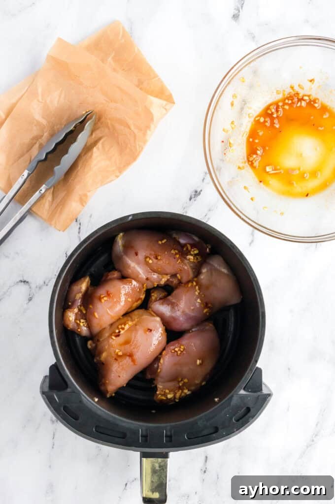 Sticky Air Fryer Chicken Thighs (Oven Method Included) 6 Marinated chicken thighs placed in a single layer in an air fryer basket on a white marble background.