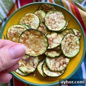 Finger holding a zucchini chip cooked in the microwave over a yellow plate with more zucchini chips.
