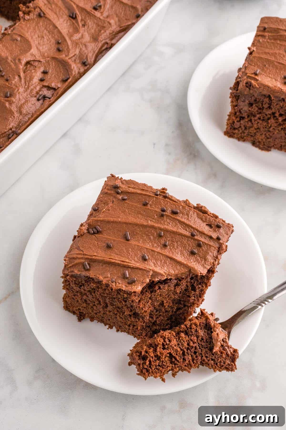 Silky Chocolate Buttercream Frosting Recipe for Cakes and Cupcakes 11 A slice of chocolate cake with chocolate buttercream on a white plate, garnished with chocolate sprinkles, next to a baking dish of more cake. A fork holds a piece of the cake.