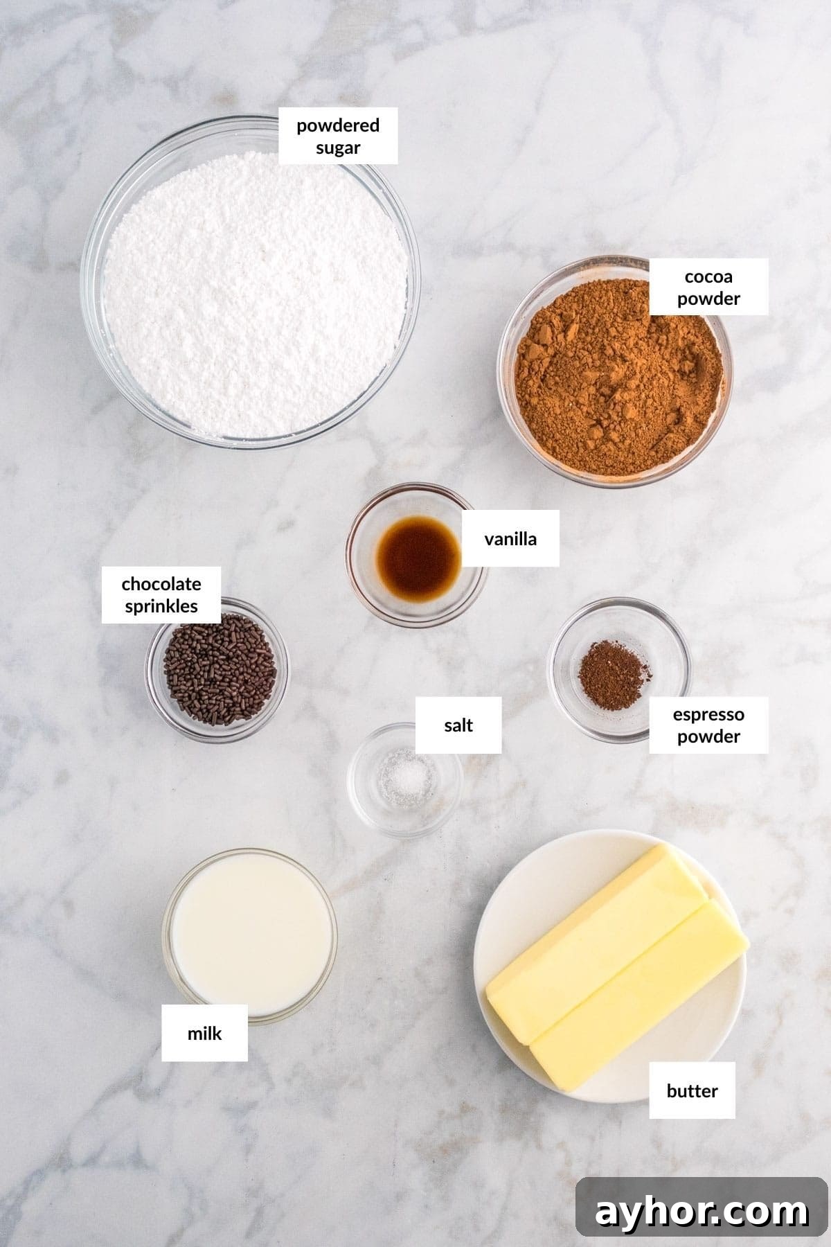 Silky Chocolate Buttercream Frosting Recipe for Cakes and Cupcakes 3 A top-down view of baking ingredients on a marble surface showcases essentials for chocolate buttercream, featuring powdered sugar, cocoa powder, vanilla, chocolate sprinkles, salt, espresso powder, milk, and butter.