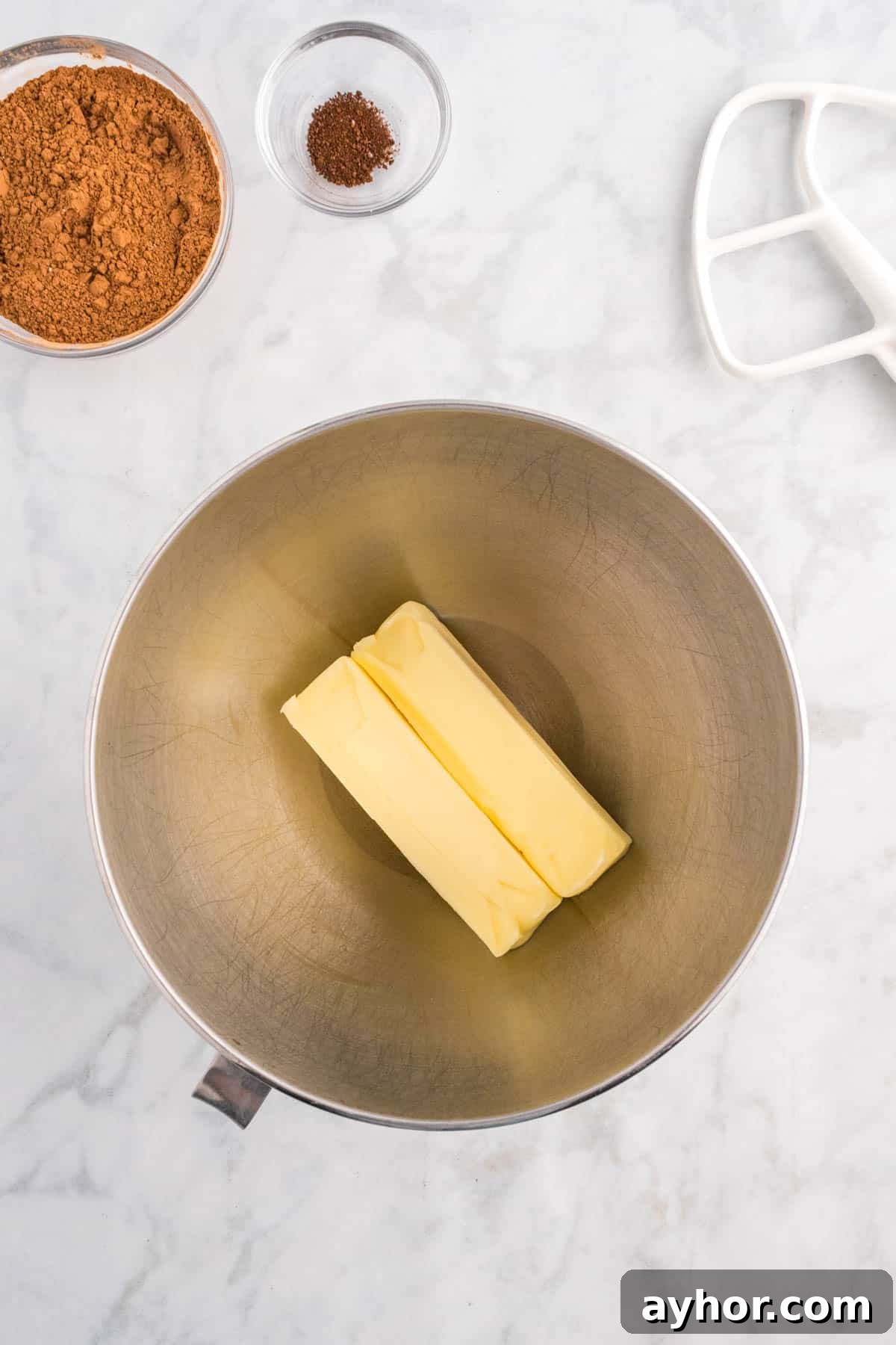 Silky Chocolate Buttercream Frosting Recipe for Cakes and Cupcakes 4 Two sticks of butter in a mixing bowl, with cocoa powder and spices in small bowls nearby on a marble countertop. A mixer attachment is visible.