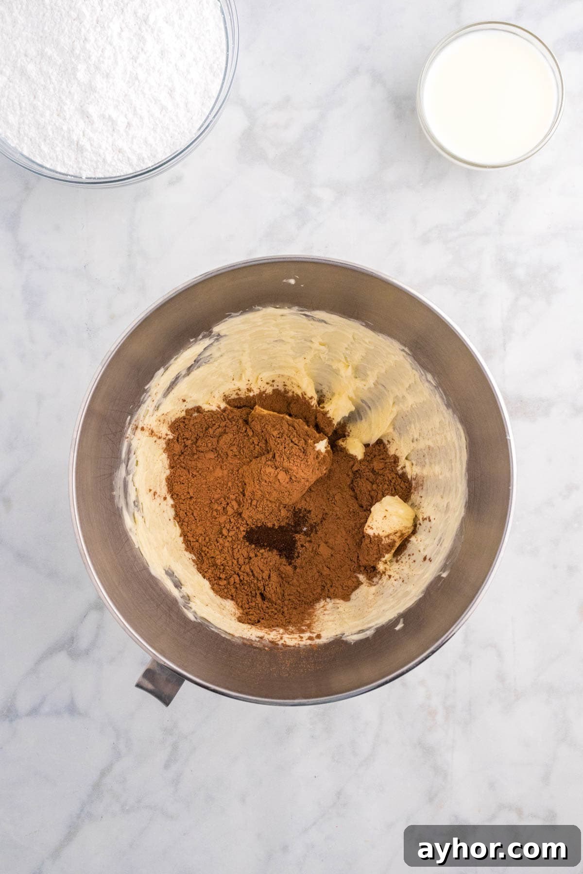 Silky Chocolate Buttercream Frosting Recipe for Cakes and Cupcakes 5 A mixing bowl contains butter, sugar, and cocoa powder on a marble countertop. Nearby are bowls of flour and a glass of milk.