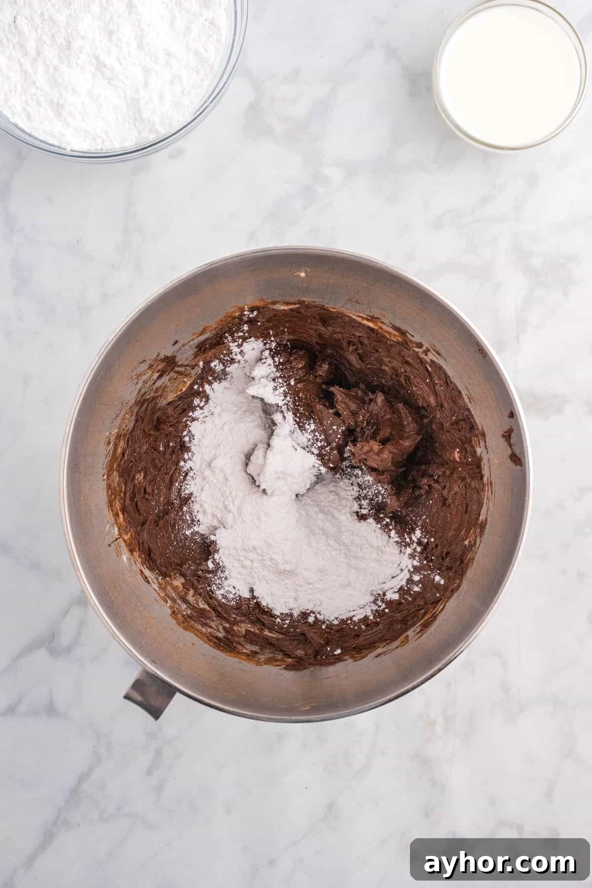 Silky Chocolate Buttercream Frosting Recipe for Cakes and Cupcakes 6 Bowl with chocolate batter and flour added on top, placed on a marble surface. Nearby, a bowl of flour and a glass of milk.