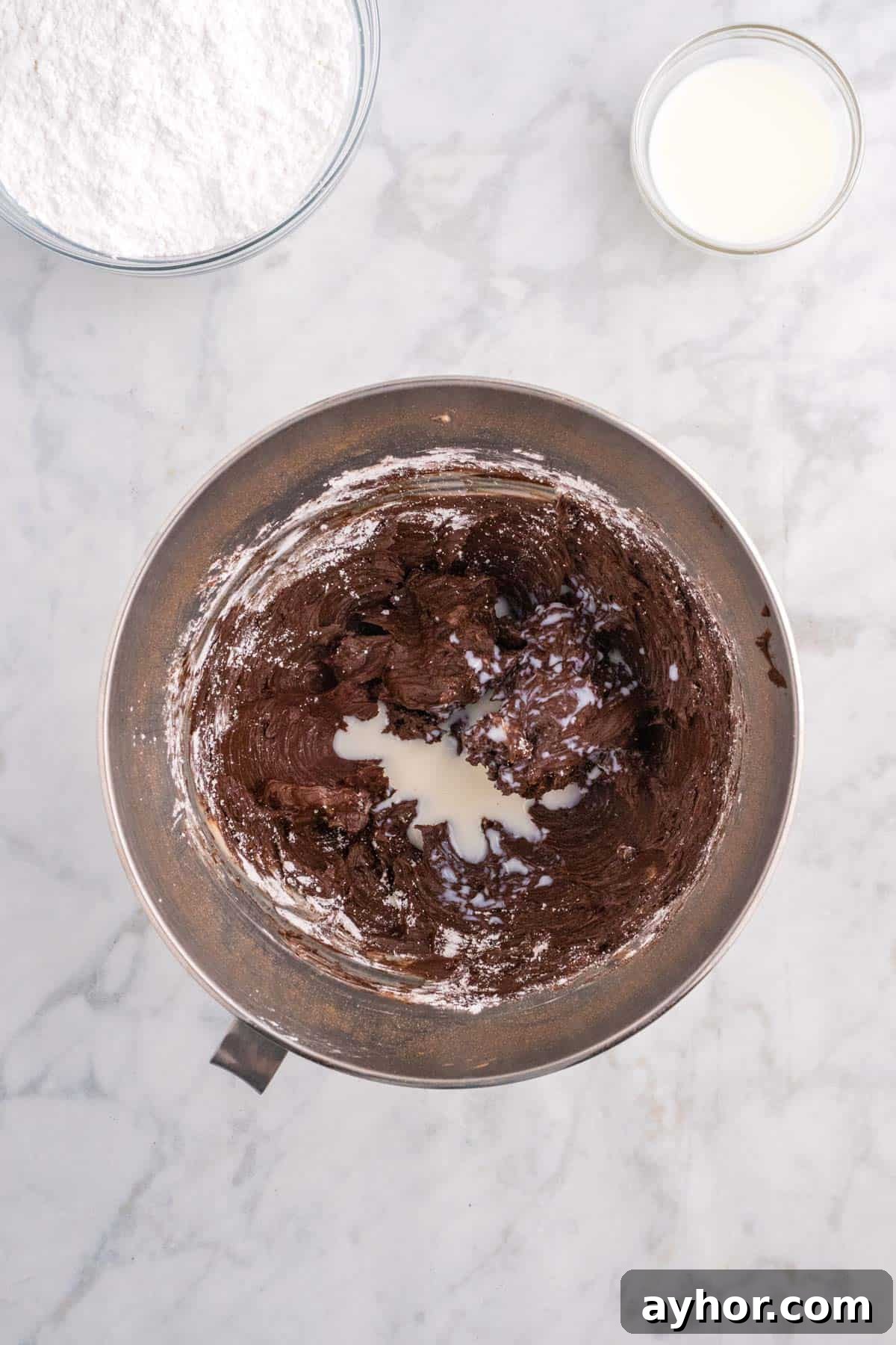 Silky Chocolate Buttercream Frosting Recipe for Cakes and Cupcakes 7 Bowl with chocolate batter being mixed, surrounded by flour in a separate bowl and a small cup of milk on a marble countertop.