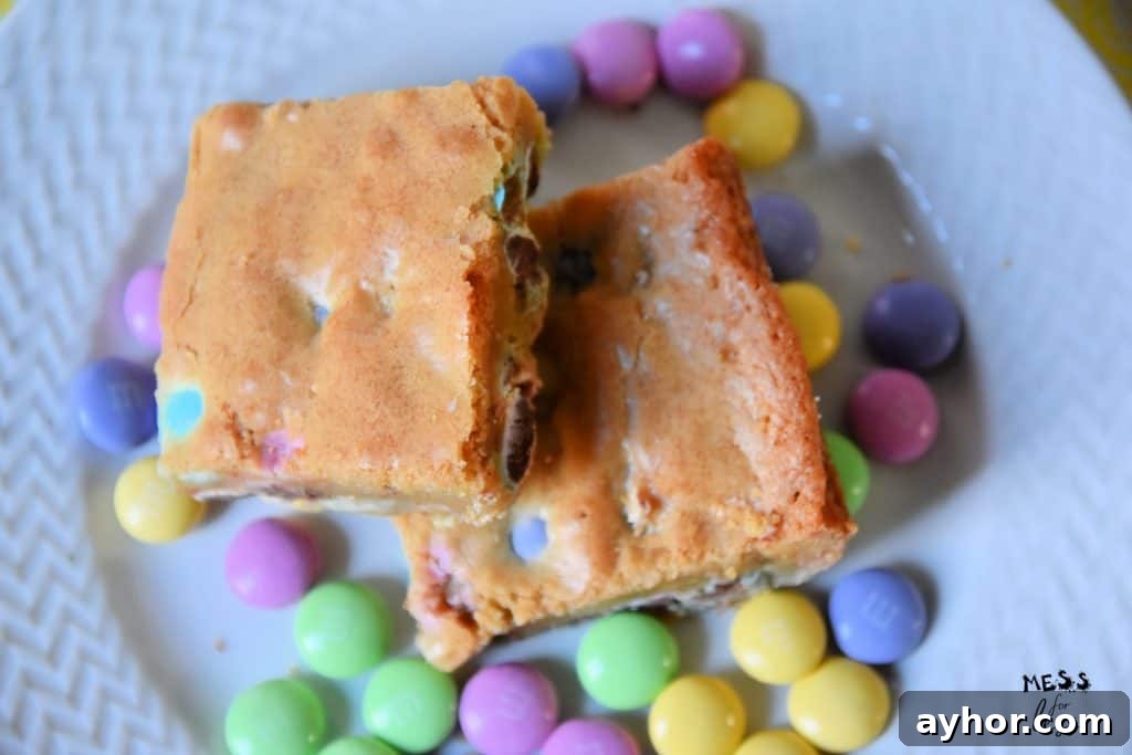 Easter Blondie Bars with Marshmallow Eggs Recipe 3 Two Easter blondies with chocolate chips rest on a plate, surrounded by colorful candy-coated chocolates.
