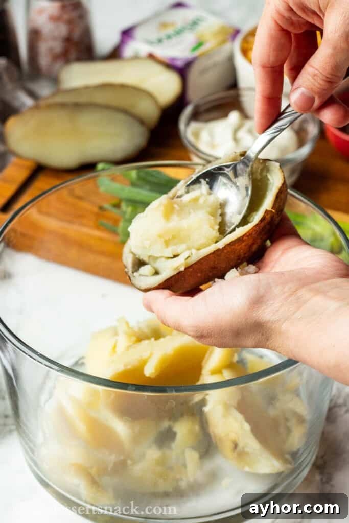 Scooping middle of baked potato with spoon into glass bowl.