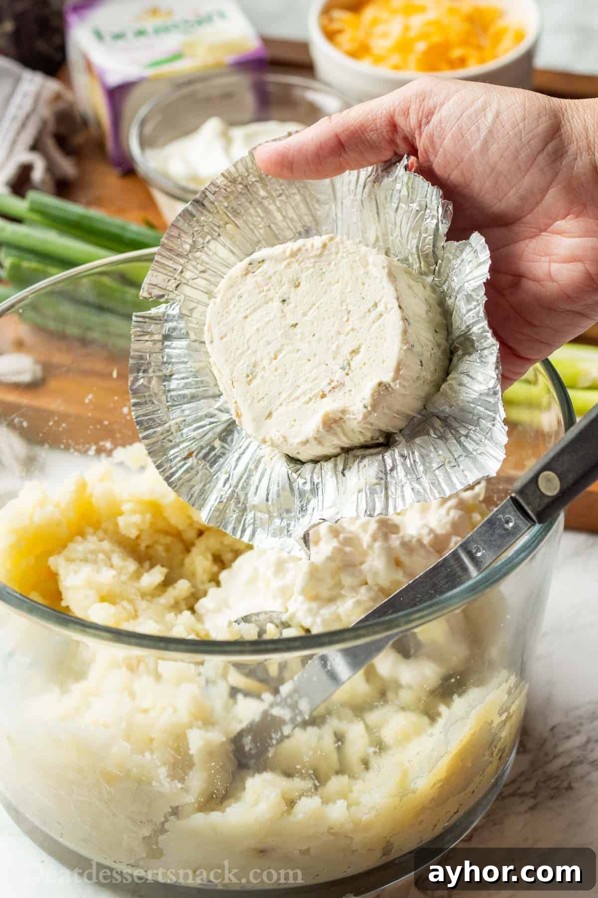 Hand holding Boursin cheese above glass bowl of potatoes.