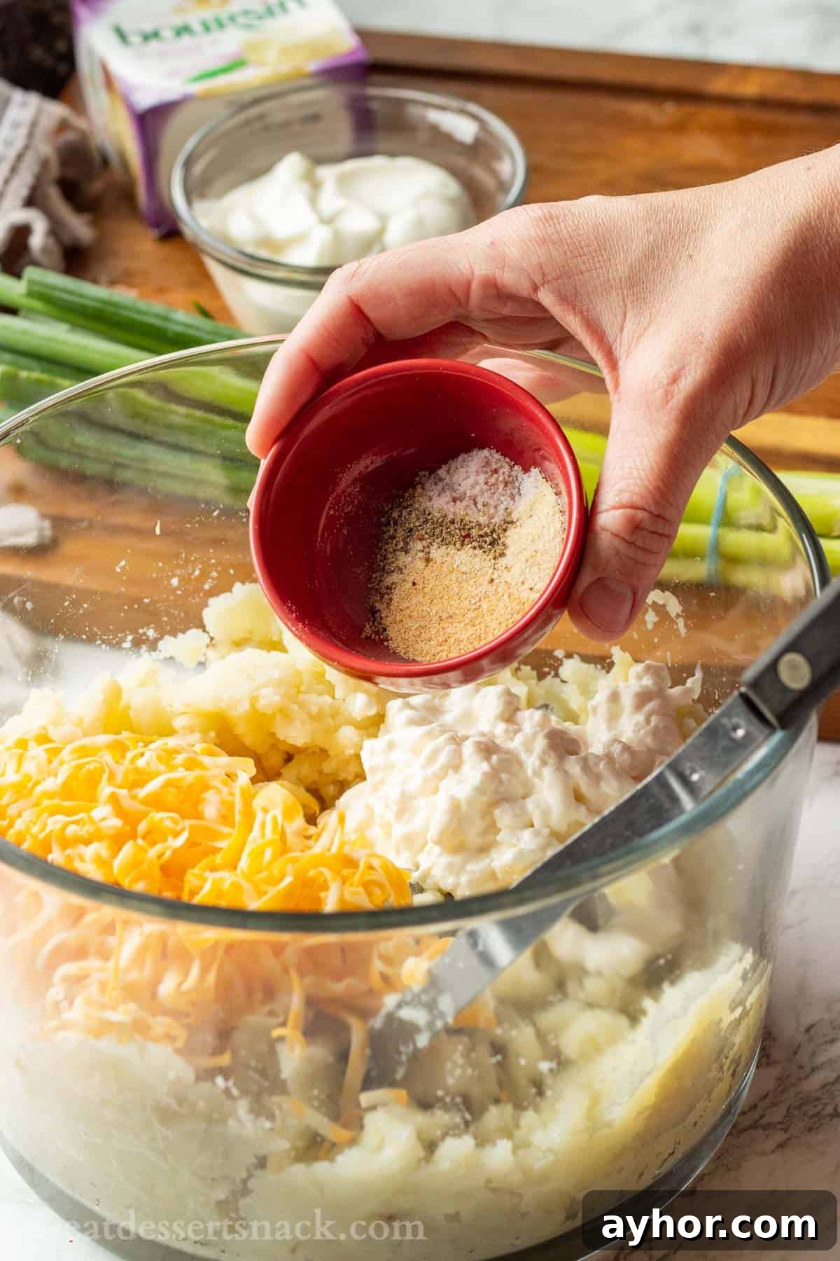 Hand pouring red bowl into glass bowl of potatoes and cheese.