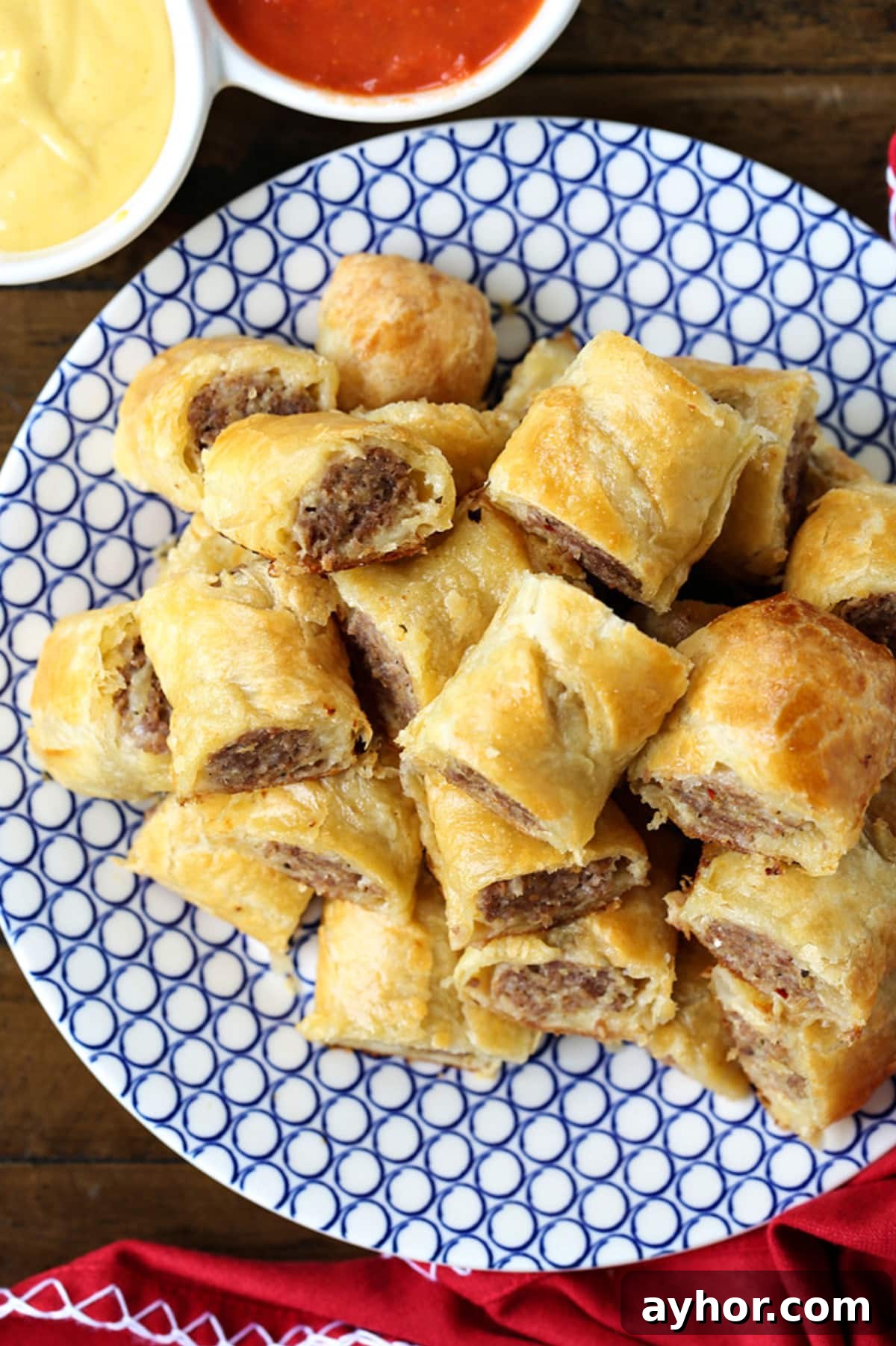 Everyone loves Puff Pastry Sausage Rolls! A buttery, flaky crust with a cheesy sausage filling. Perfect for holidays, parties and game days! sausage rolls stacked on plate with ketchup and mustard