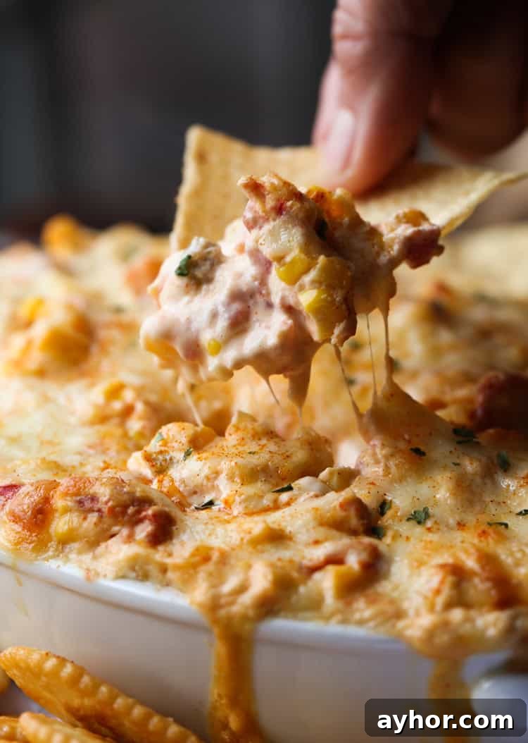 Warm and creamy hot corn dip is the ultimate party appetizer! It's baked until bubbly and delicious with chips, crackers, or veggies. A hand lifting a corn chip from hot corn dip in a casserole dish.
