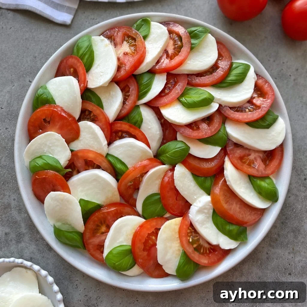 Classic Tomato Caprese Salad with Fresh Basil and Mozzarella 2 A plate of tomato caprese salad arranged in a circular pattern with alternating slices of fresh mozzarella, ripe tomatoes, and basil leaves on a gray surface.