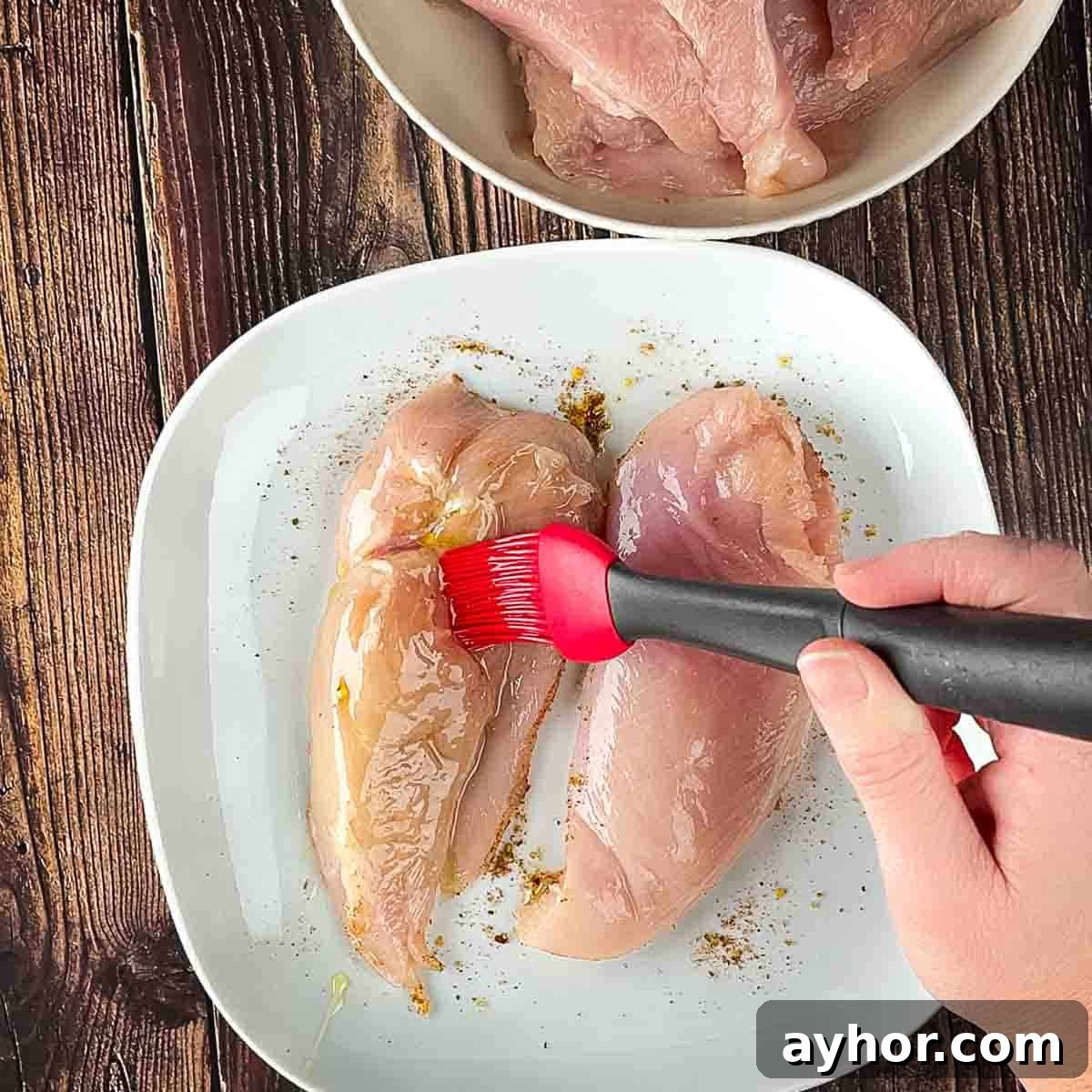 A red basting brush adding oil to the top of the chicken breast.