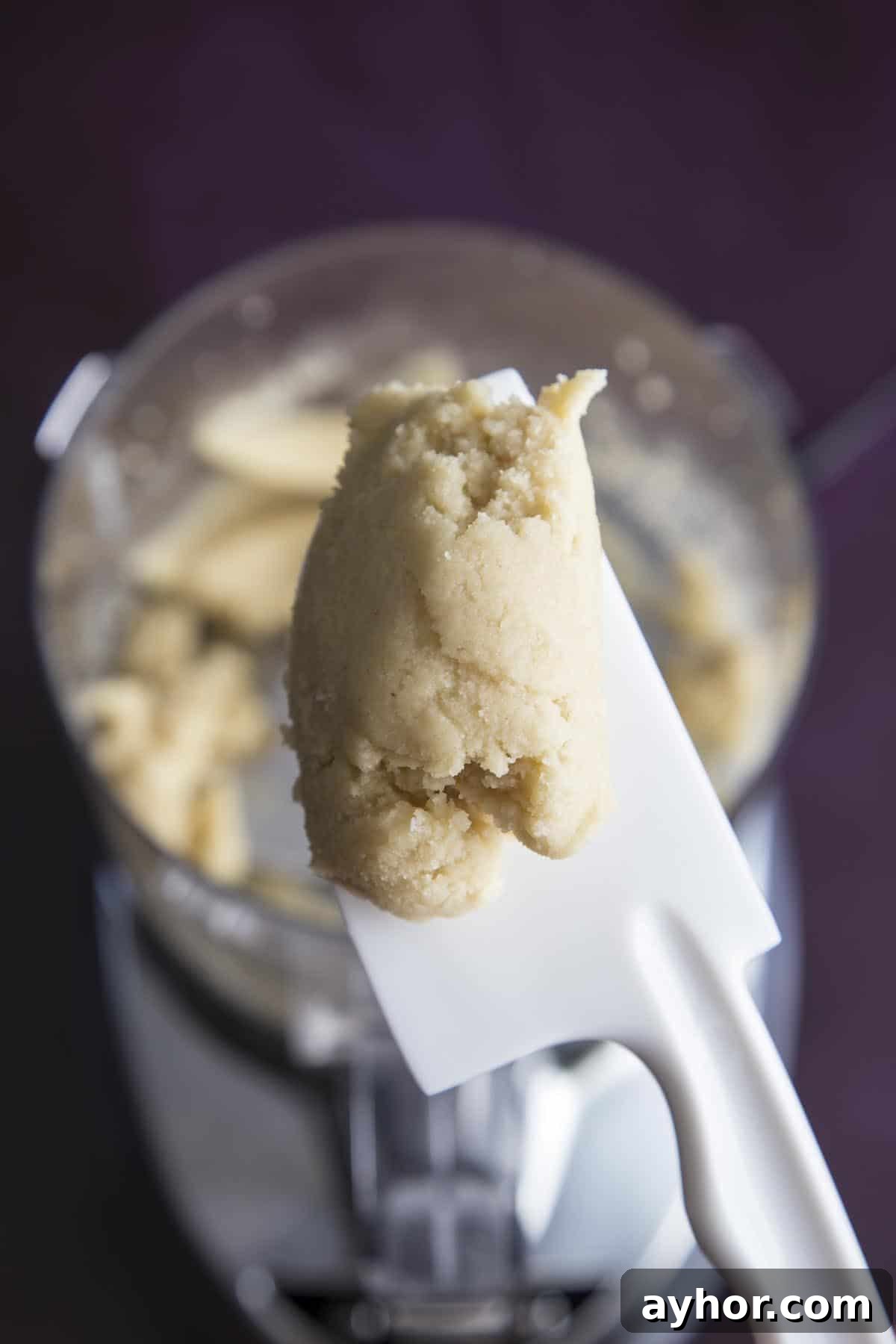Homemade Almond Paste can be made in as little as 5 minutes with just 5 ingredients. It is a versatile and delicious filling that will elevate your almond desserts to the next level. #wildwildwhisk #almondpaste A spatula holding a piece of almond paste just made in a food processor.
