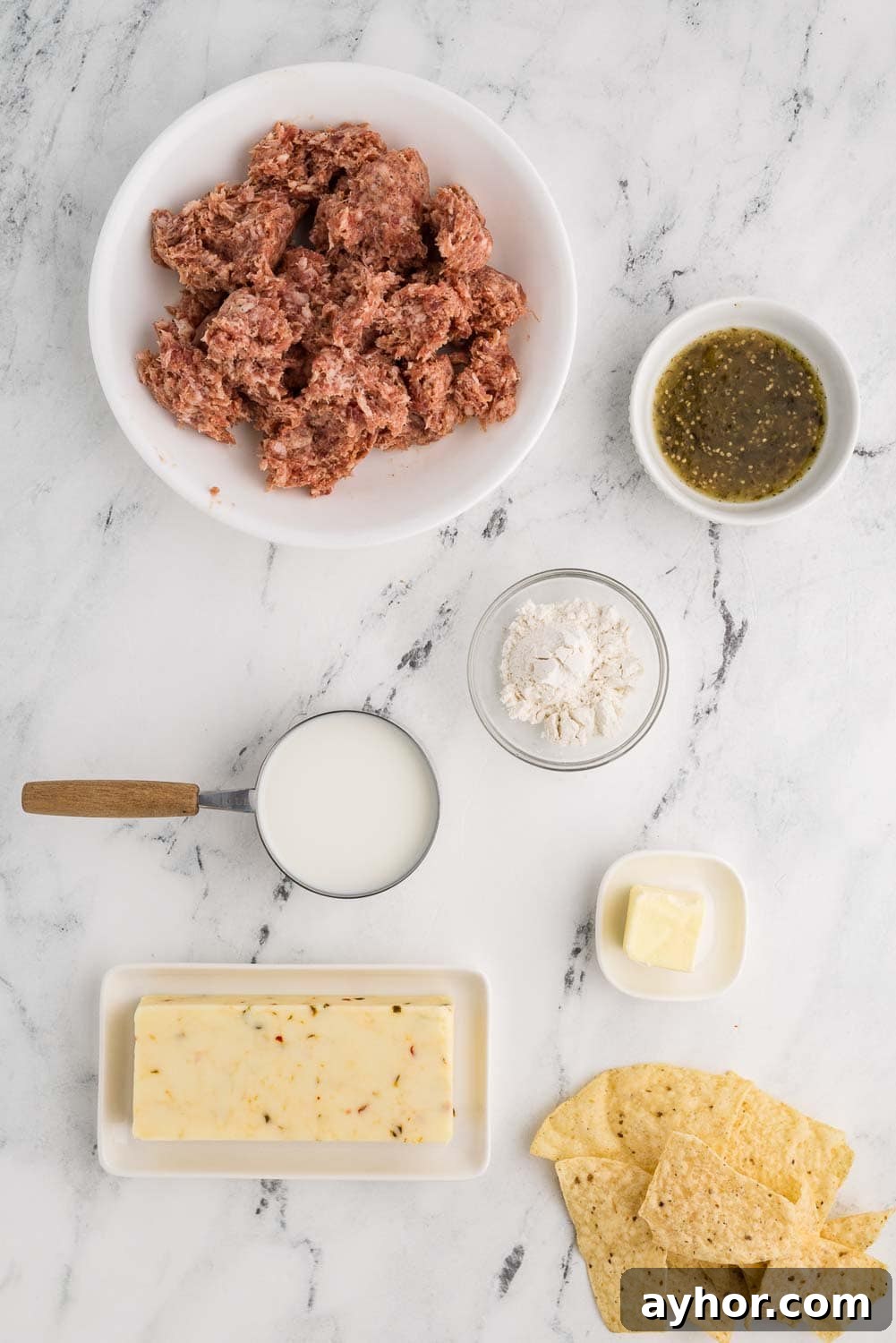 Ingredients for a sausage cheese dip recipe arranged on a kitchen counter. Includes 12 oz of ground pork sausage, an 8 oz block of pepper jack cheese, a small bowl with 2 tablespoons of flour, a stick of butter, a measuring cup with 1 cup of milk, a small bowl of salsa verde, and tortilla chips laid on counter
