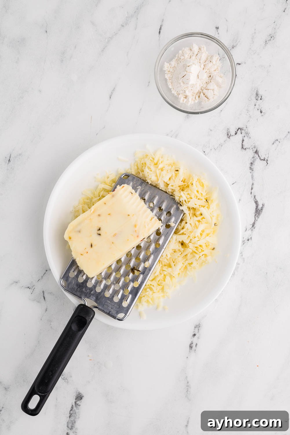 : A bowl filled with shredded pepper jack cheese, with a box grater laid across the top. A partially used block of pepper jack cheese rests on the grater, ready to be shredded further.