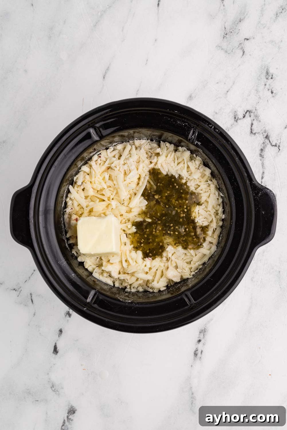 Ingredients for sausage queso dip inside a slow cooker. Includes cooked ground pork sausage, shredded pepper jack cheese, flour, butter, milk, and salsa verde, layered and ready to cook.