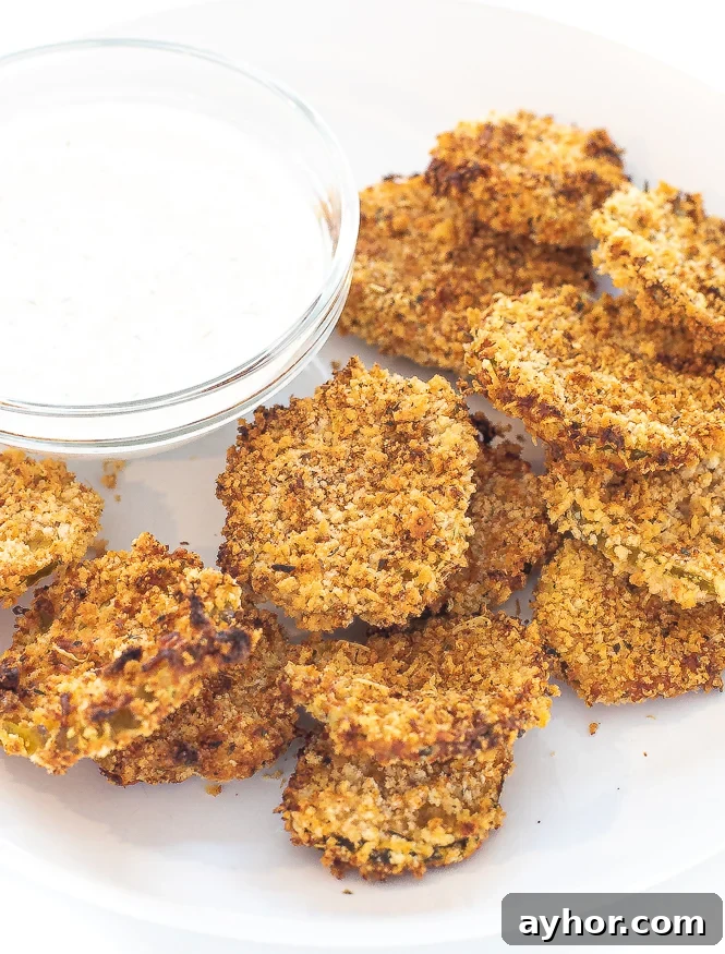 Crispy Air-Fryer Pickles: Tangy Homemade Fried Pickle Chips 2 Air fryer pickle chips stacked on a white plate next to a bowl of sauce.