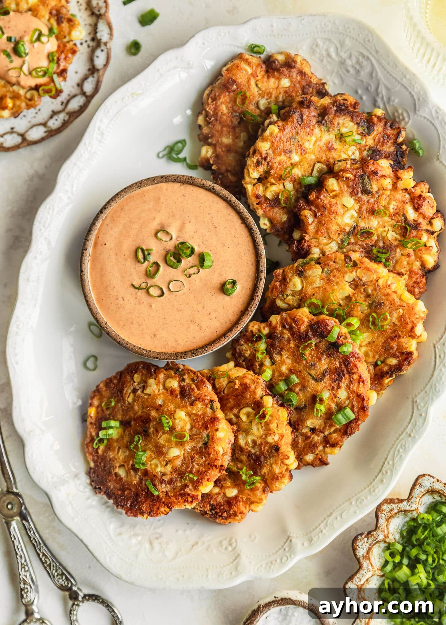 A white platter of bacon and corn fritters with a brown bowl of chipotle aioli on a beige counter next to silver tongs, a glass of white wine, and white and brown bowls of salt and green onions. 