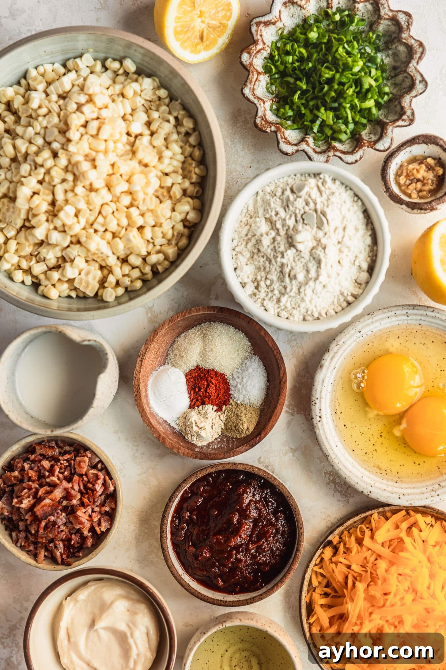 White and brown bowls of corn, green onions, flour, garlic, milk, spices, eggs, bacon, cheese, chipotle peppers, mayo, oil, and lemons on a beige counter.