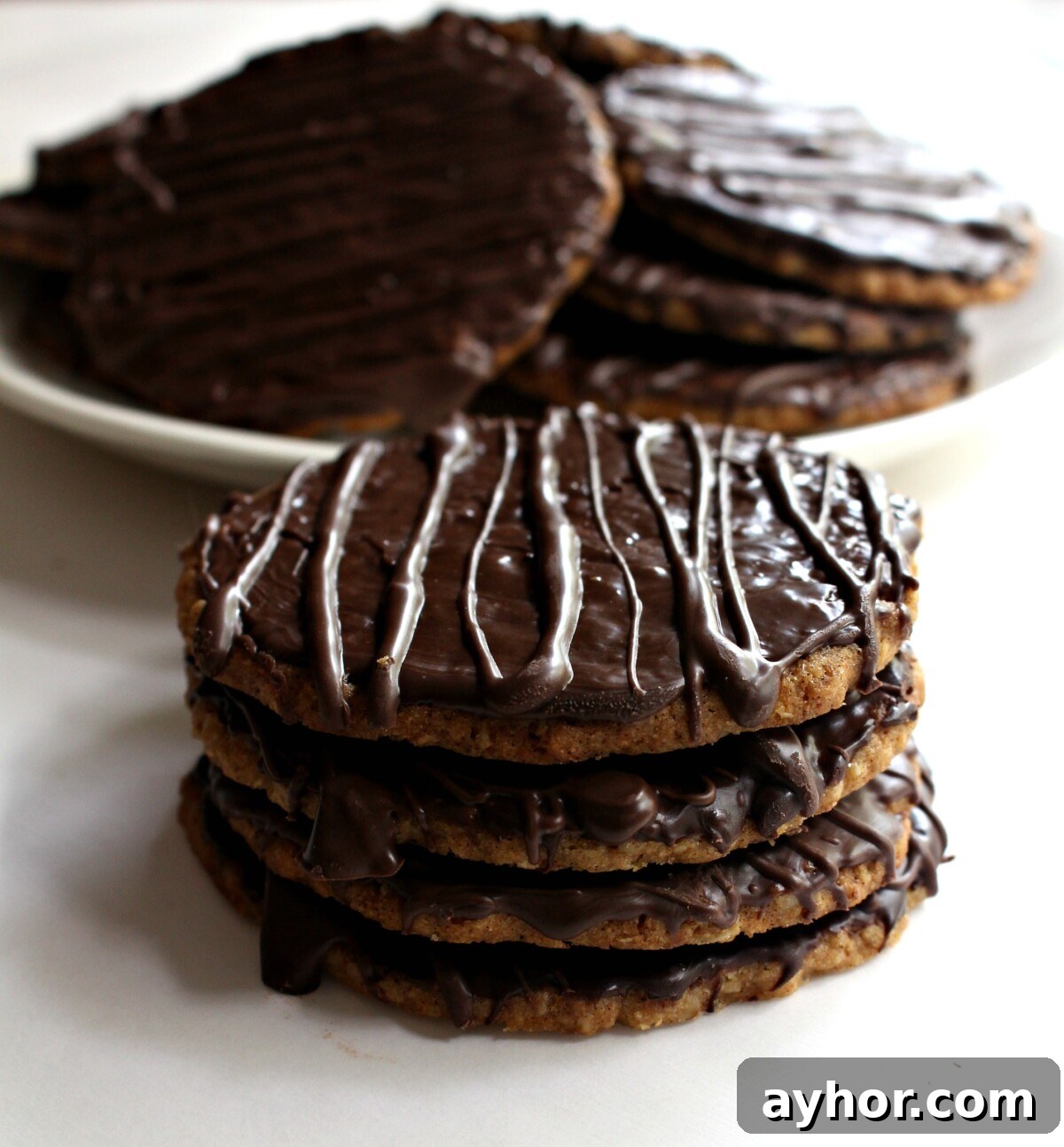 Chocolate-Covered Hobnob Oat Cookies Recipe 4 Four cookies in a stack and a white plate of cookies in the background.