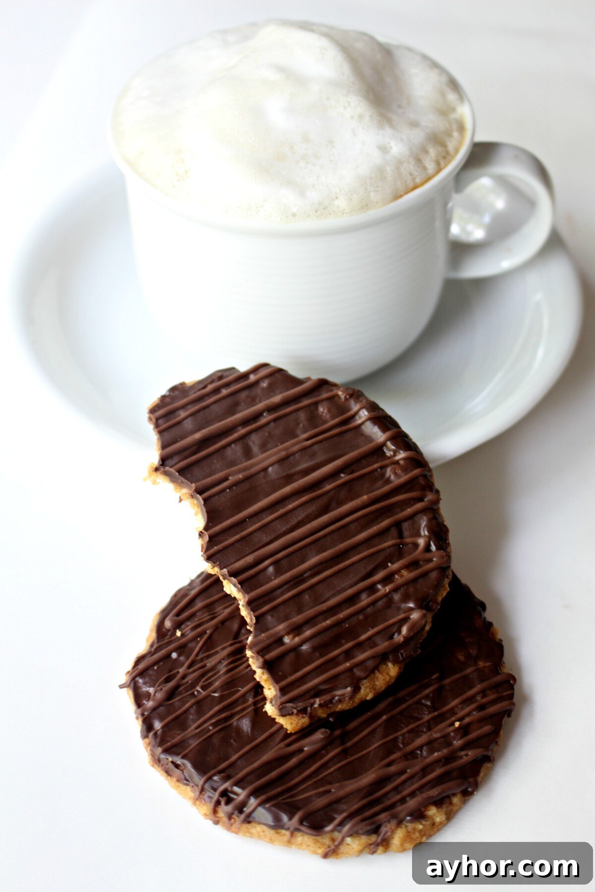 Chocolate-Covered Hobnob Oat Cookies Recipe 8 Two cookies in front of a white mug of cappuccino topped with milk foam.