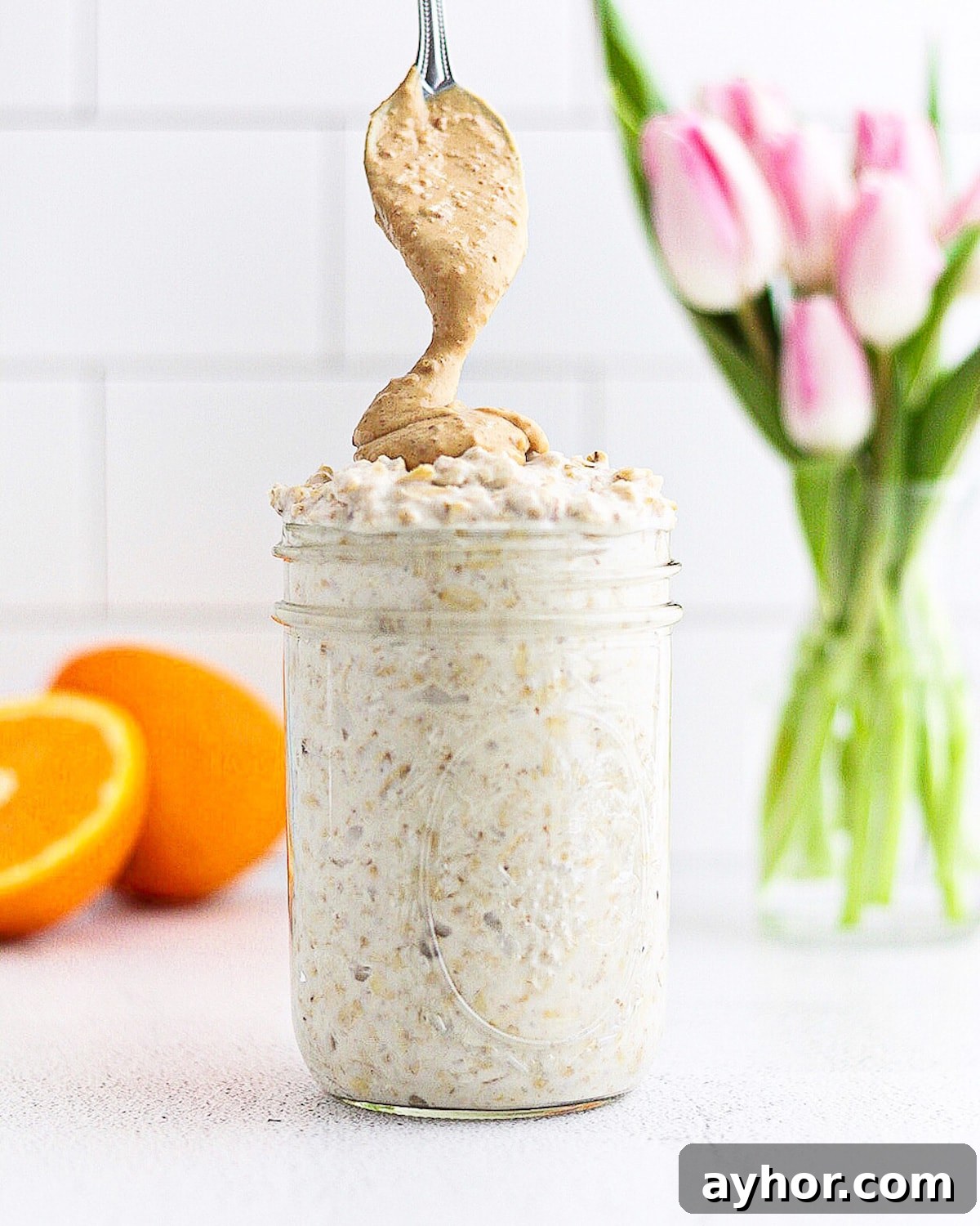 Creamy Overnight Oats with Fruit and Nuts — Make Ahead Breakfast 2 A mason jar with overnight oats inside. There is a silver spoon drizzling peanut butter on top of the oats. There is an orange and a vase of pink tulips in the back.