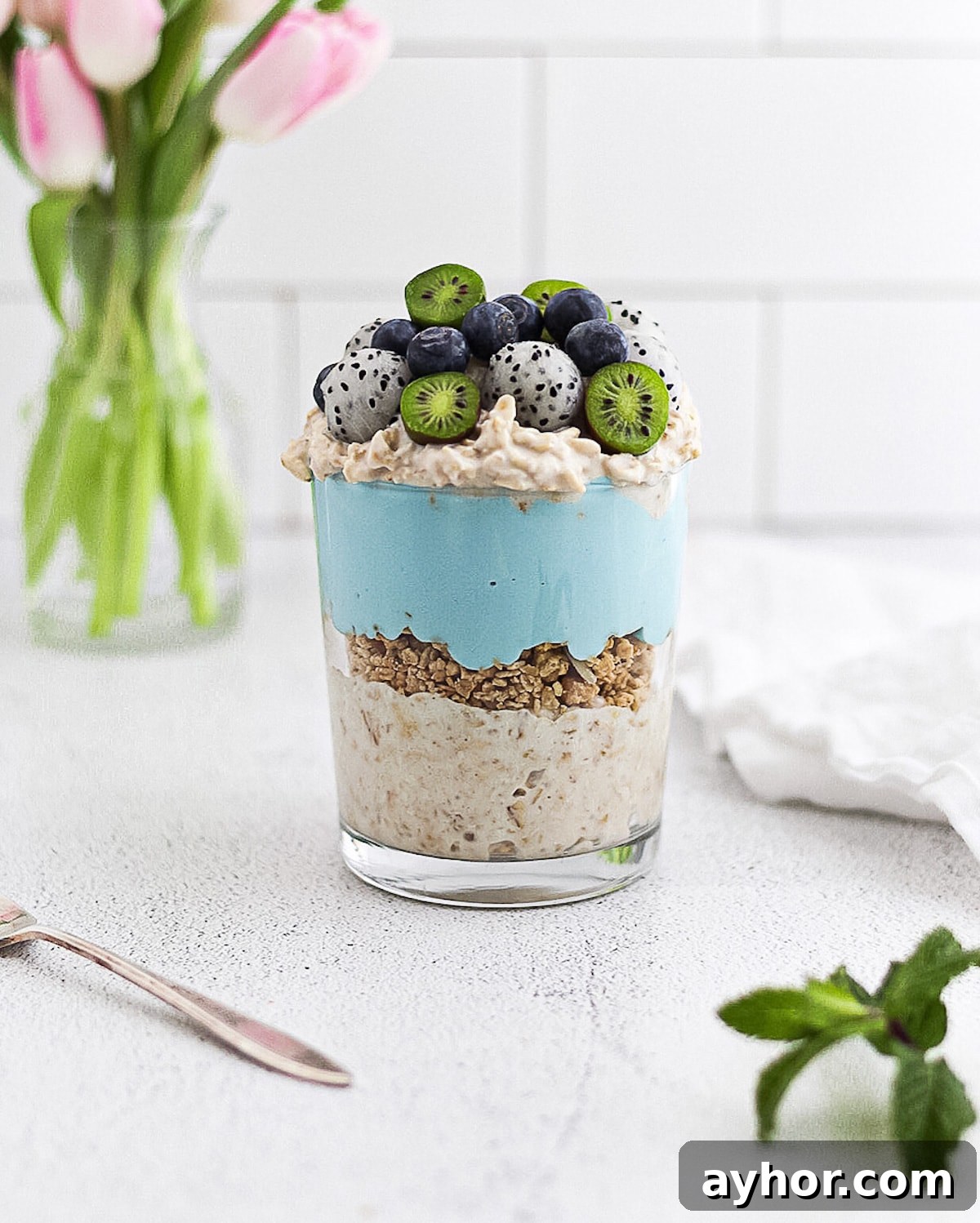Creamy Overnight Oats with Fruit and Nuts — Make Ahead Breakfast 5 A clear glass filled with a parfait including a layer of overnight oatmeal, granola, blue spirulina yogurt, overnight oats, and a mixture of fresh fruit on top; kiwi berries, blueberries, and dragonfruit balls.
