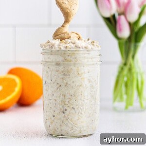 Creamy Overnight Oats with Fruit and Nuts — Make Ahead Breakfast 6 A picture of a clear mason jar with overnight oatmeal inside. A spoon is drizzling peanut butter on top. There are pink tulips and a sliced orange in the background.