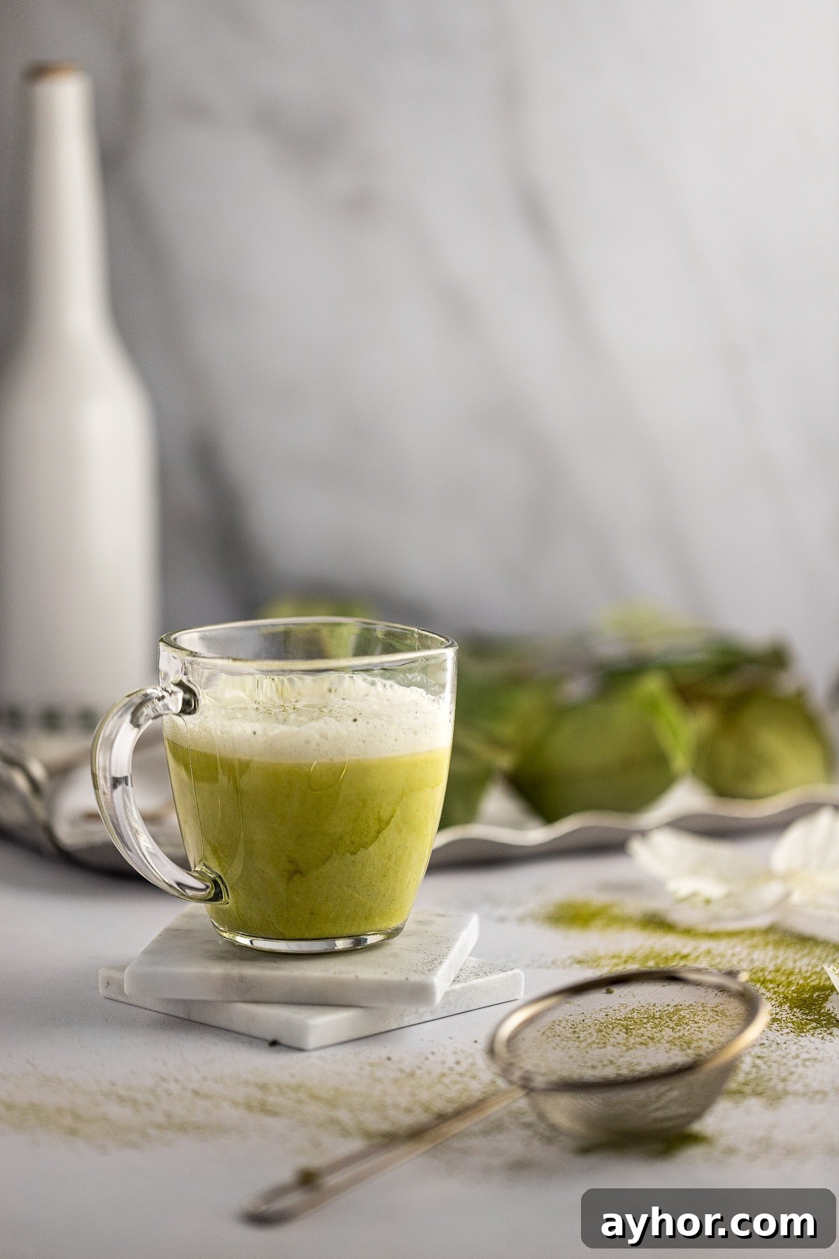 A glass of almond milk matcha latte on two white marble coasters, with green plants in the background and a metal strainer in the foreground.
