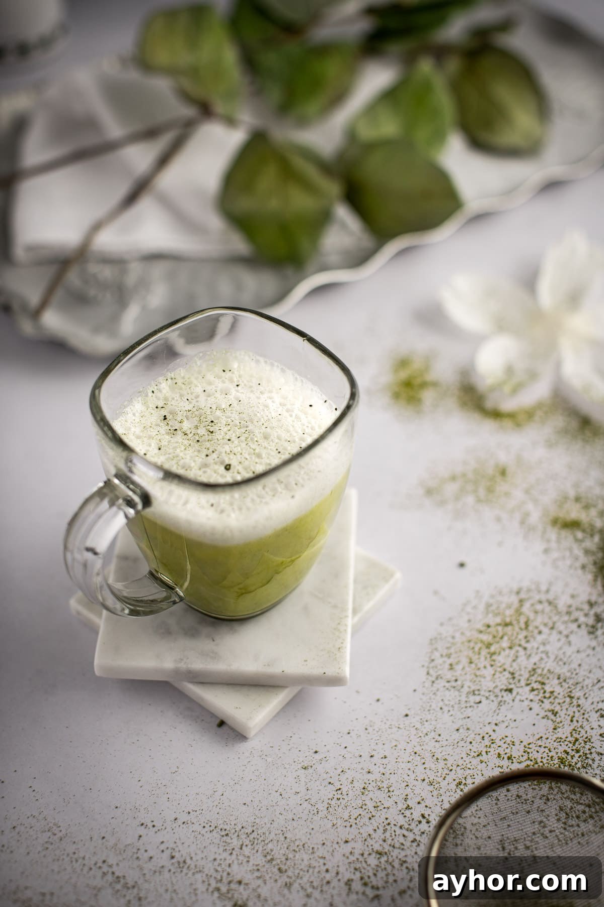 Matcha almond milk latte on two white marble coaster, on a light grey marble table, with white flowers and matcha powder scattered around.