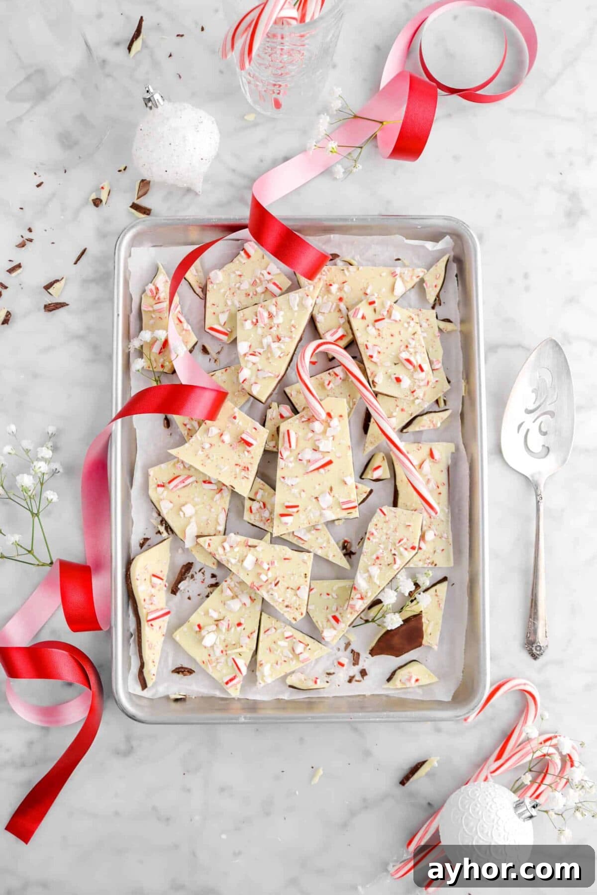 peppermint bark on sheet pan with red ribbon, white ornaments, white flowers, and candy canes