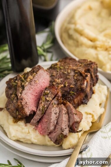 Sliced medium-rare lamb loin chops served on a bed of mashed potatoes, garnished with herbs, with a fork on the plate and a bowl of mashed potatoes in the background.