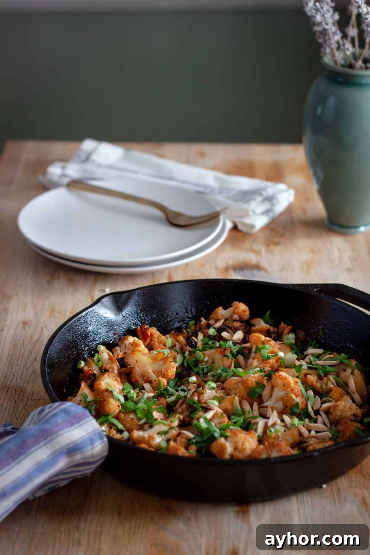 A cast iron skillet with sweet chili roasted cauliflower topped with parsley, almonds and raisins on a butcherblock countertop.