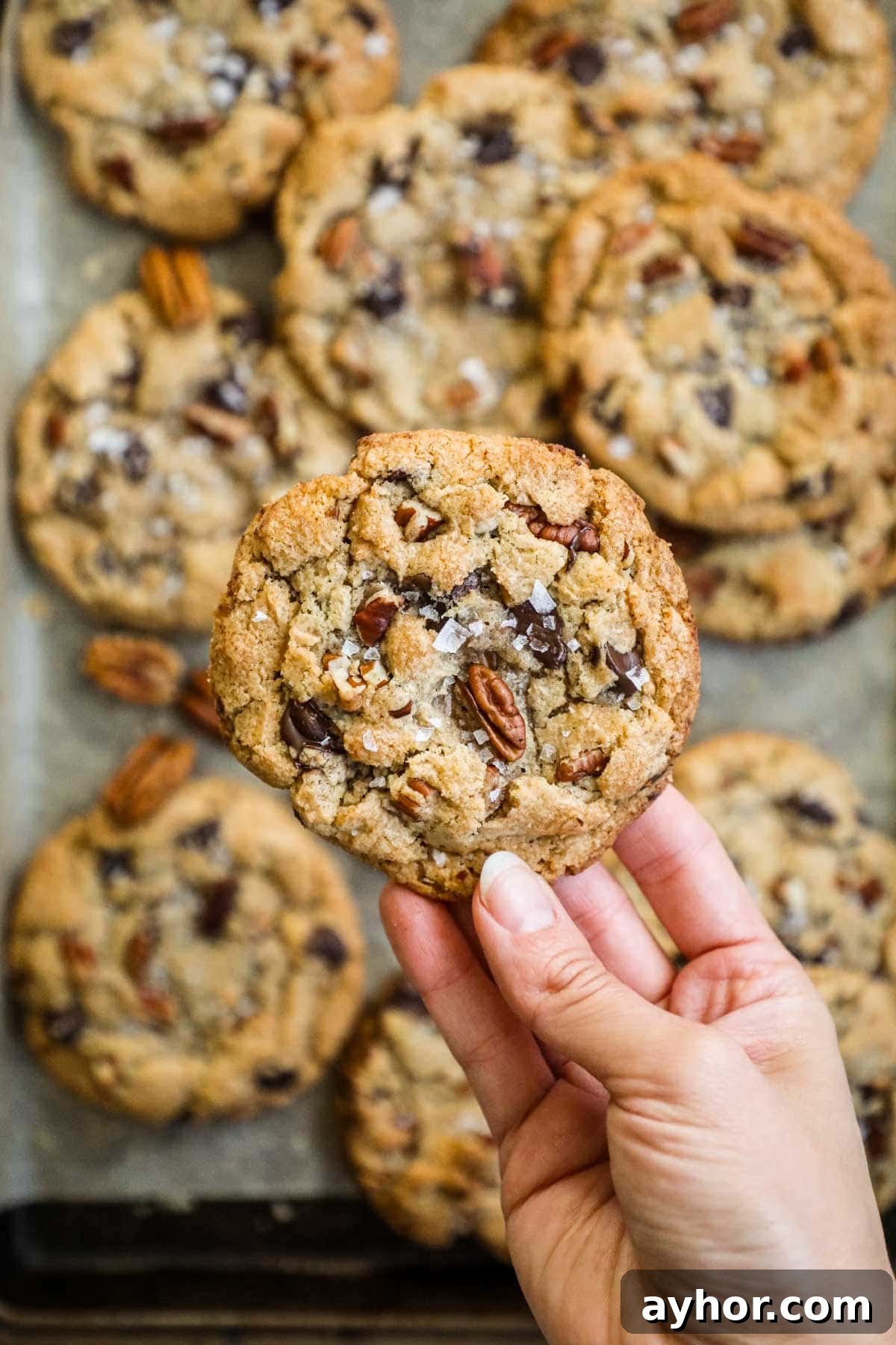 Hand holding a browned butter pecan chocolate chip cookie up close above a tray of cookies.