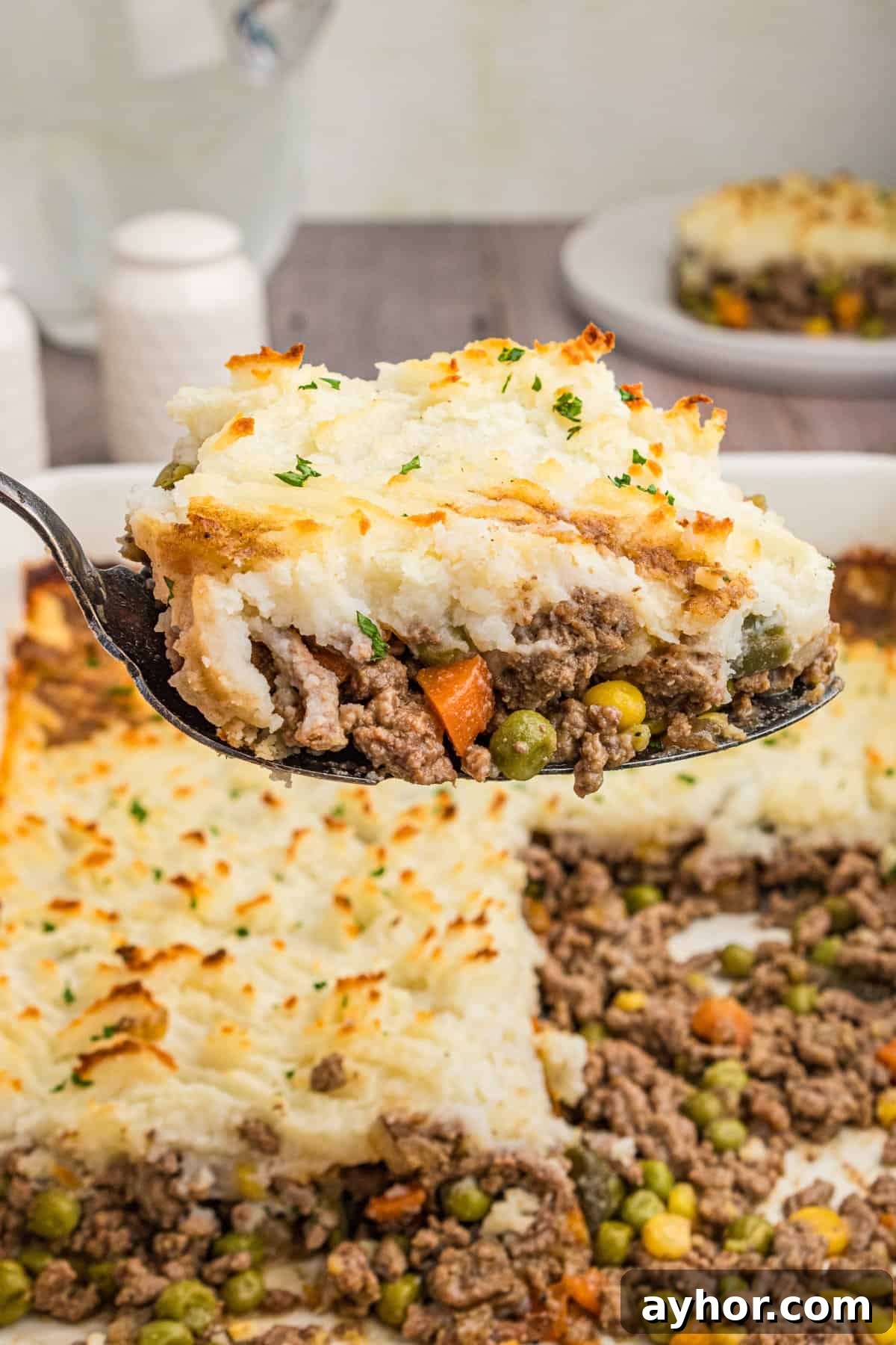 A close-up of a spoonful of cottage pie, showing golden mashed potatoes on top of a beef and vegetable mixture, garnished with chopped parsley. The casserole dish with the remaining pie is visible in the background.