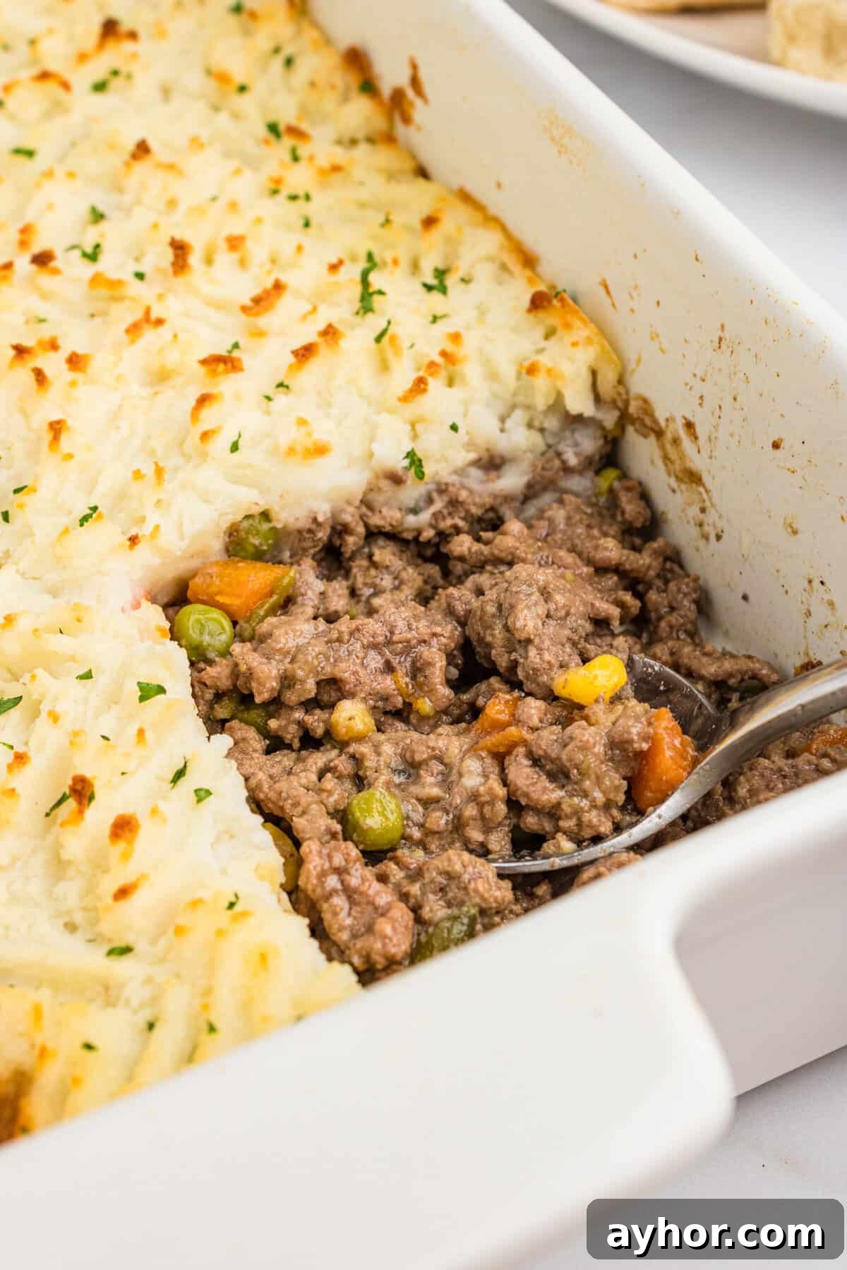 A close-up of a white casserole dish with a portion of cottage pie scooped out, revealing the seasoned ground beef, peas, carrots, corn, and green beans beneath the golden mashed potato topping.