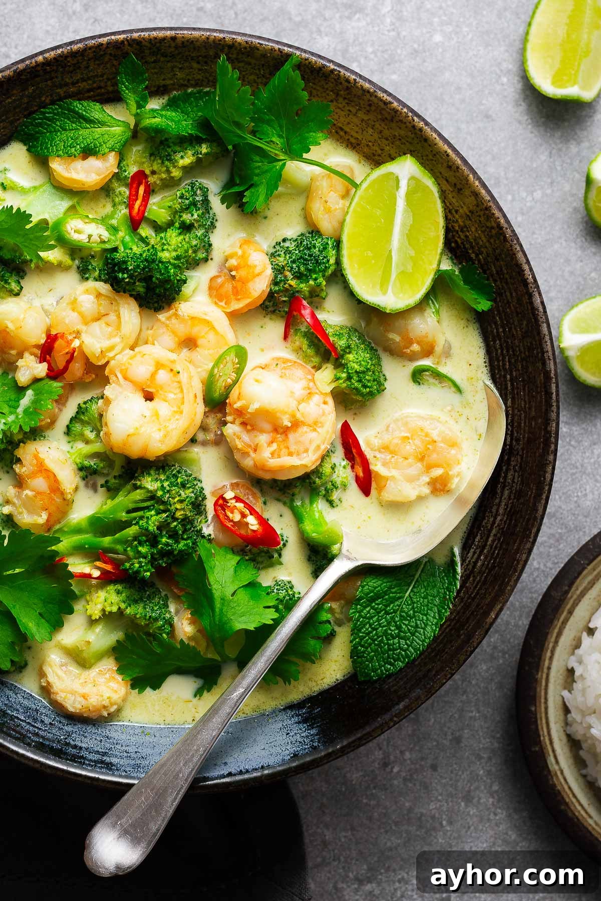 A ceramic bowl with Thai green curry with shrimp and broccoli garnished with fresh herbs and lime wedges with a serving spoon.