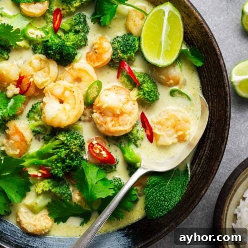 A ceramic bowl with Thai green curry with shrimp and broccoli garnished with fresh herbs and lime wedges with a serving spoon.