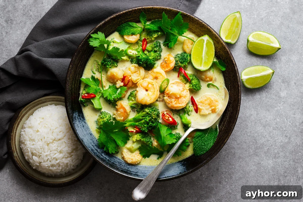 A Thai green curry bowl with shrimp and green vegetables garnished with fresh herbs and lime wedges with a serving spoon and a side or rice.