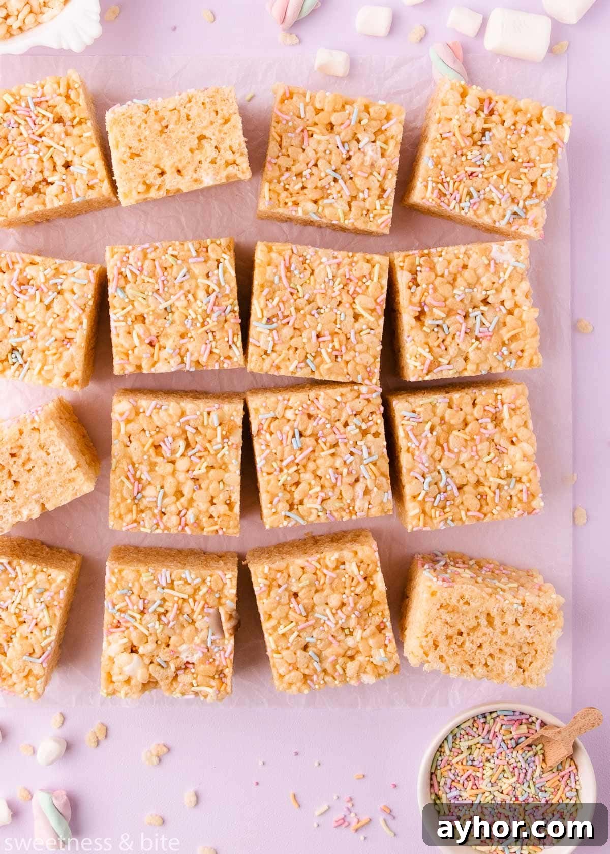 Gluten-Free Rice Krispie Treats Recipe That Really Crisps 10 Squares of gluten free rice krispie treats arranged haphazardly on a sheet of crinkled parchment on a lilac background, with mini marshmallows, rice krispies and sprinkles scattered around.