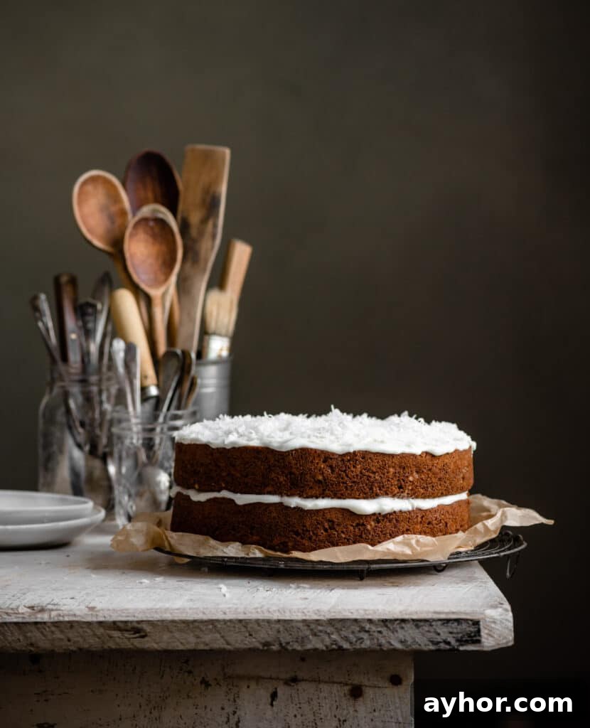 Moist Carrot Cake with Cream Cheese Frosting Recipe 11 Two layered carrot cake with coconut icing on a white table next to jars of spoons.