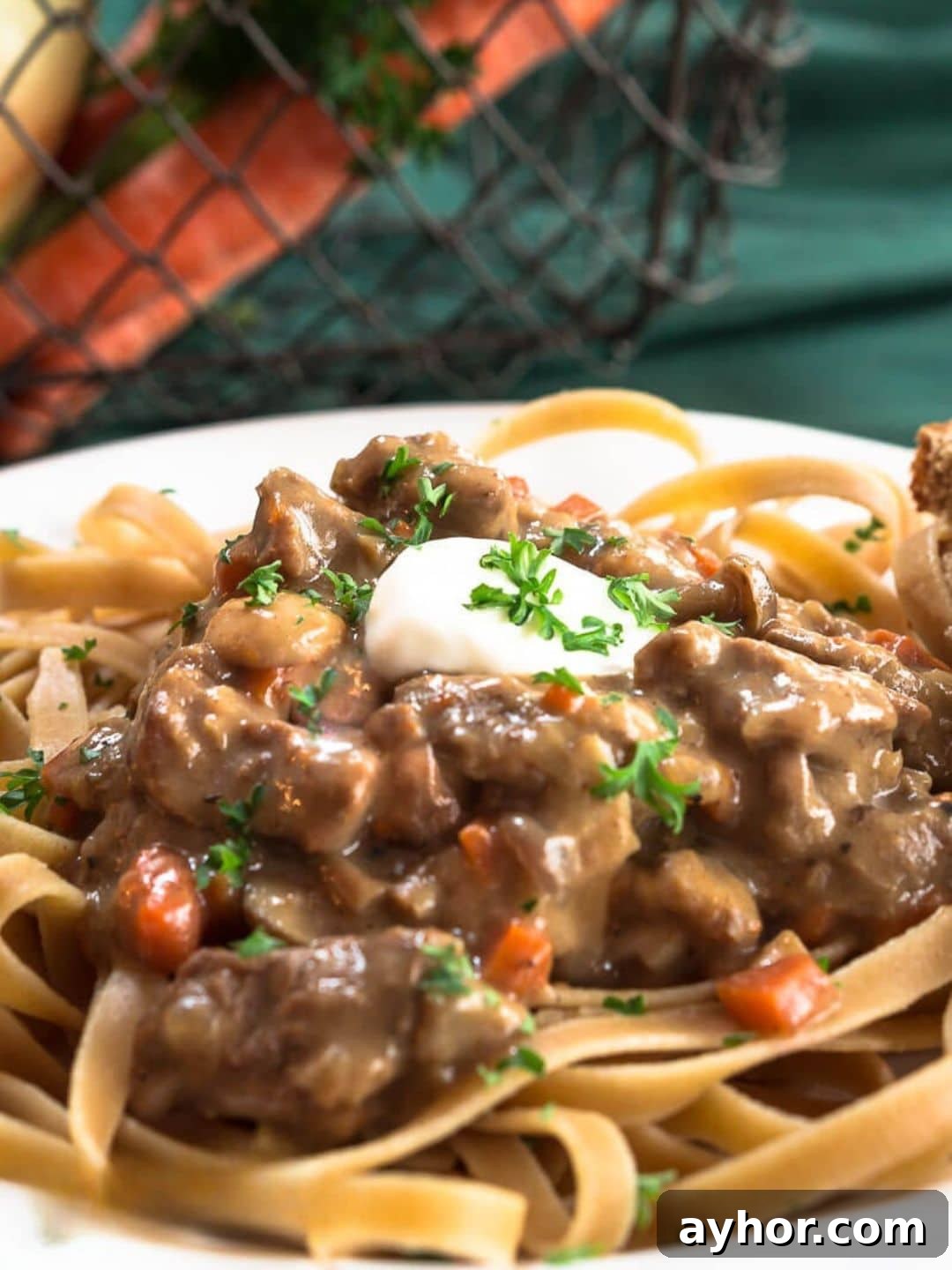 Close up view of tender chunks of beef in a stroganoff sauce served over whole grain pasta noodles and garnished with sour cream and chopped parsley - Hostess At Heart