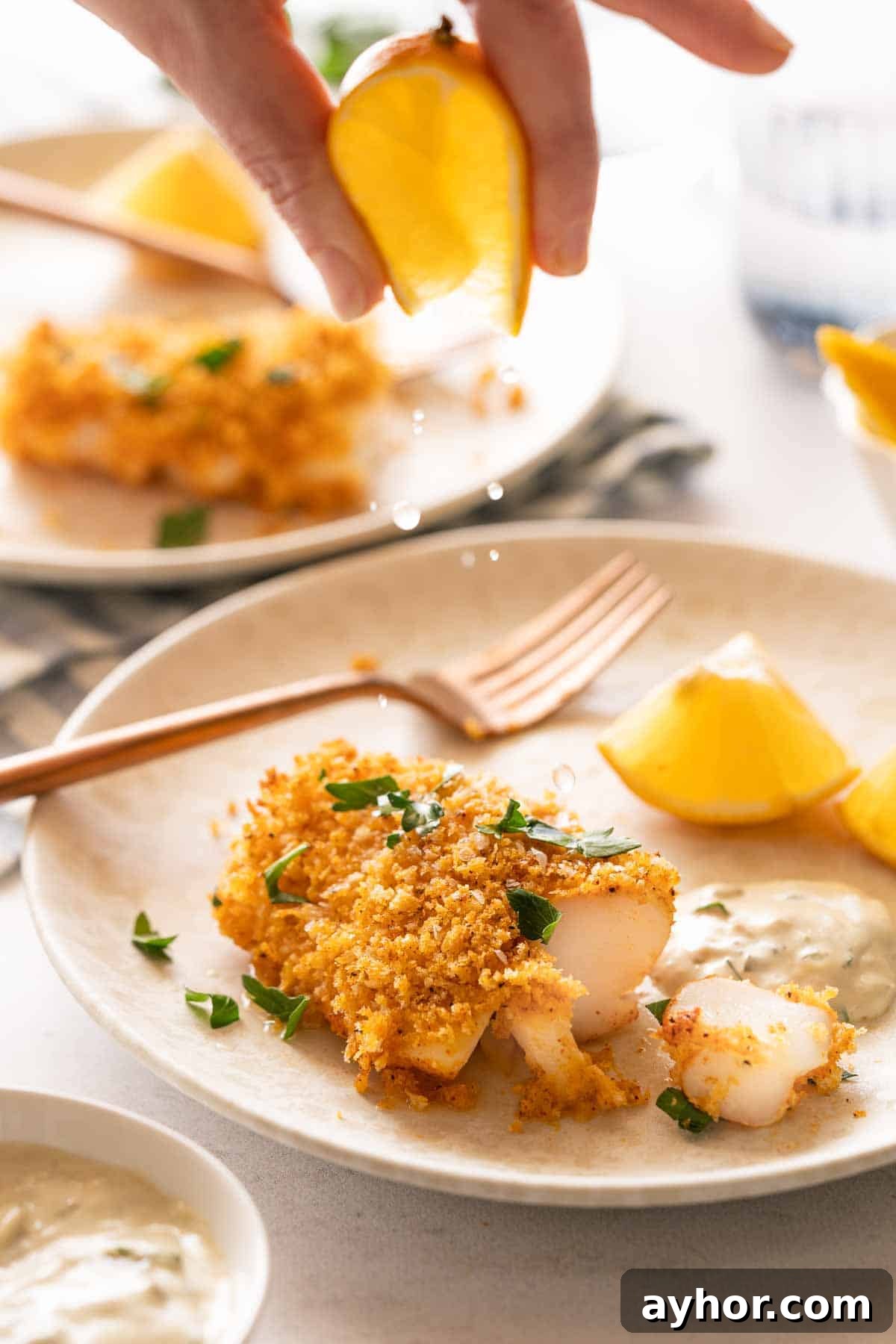 Crispy Panko-Crusted Cod with Lemon Herb Sauce 2 lemon being squeezed over panko crusted cod with tartar sauce and lemon on the side.