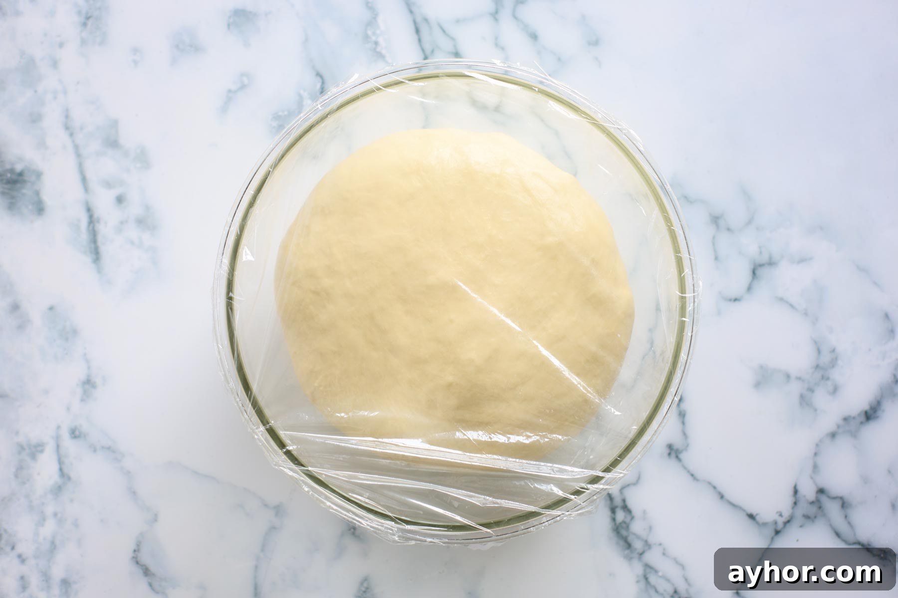 Dough after first rise, covered with plastic wrap inside glass bowl.