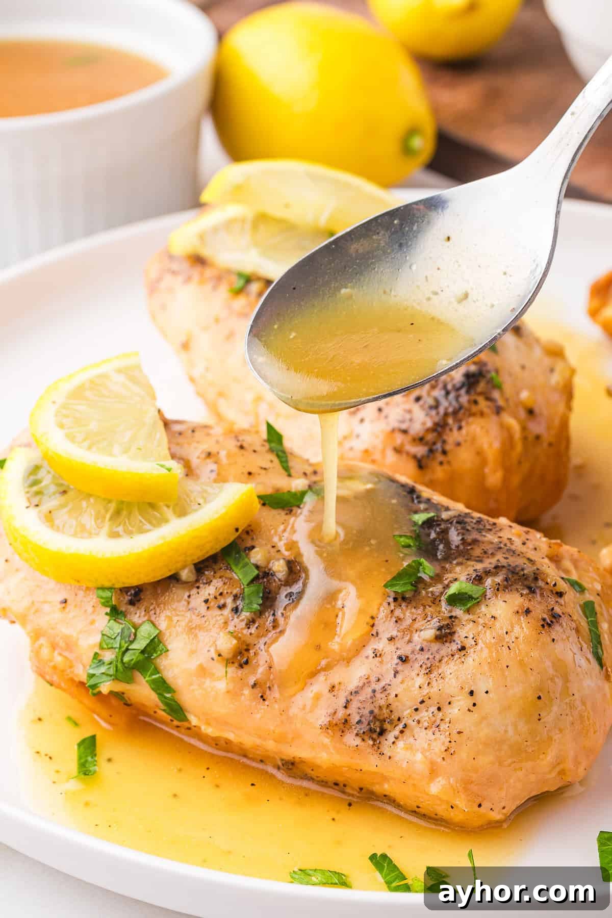 A spoon drizzles lemon sauce over seasoned Lemonade Chicken, garnished with lemon slices and herbs.