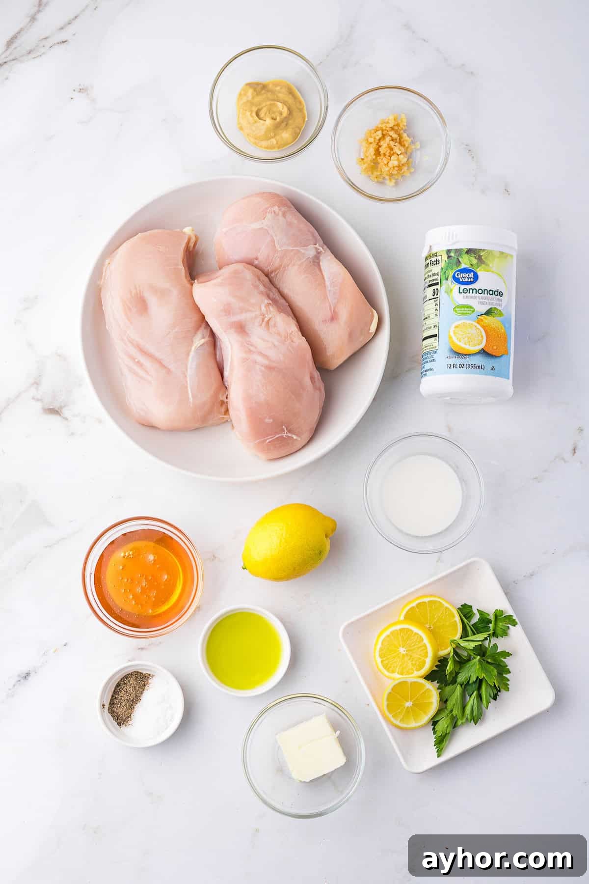 Ingredients on a marble surface: raw chicken breasts, mustard, minced garlic, lemon, honey, olive oil, butter, lemon slices, parsley, seasoning and lemonade concentrate.