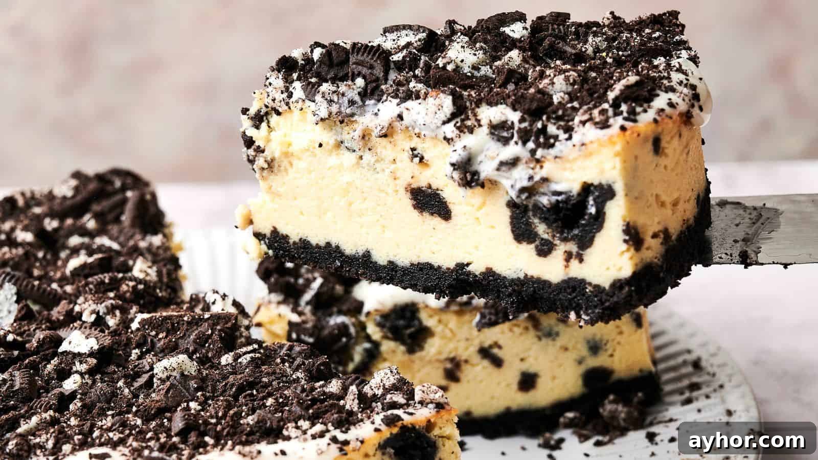 A slice of cookies and cream cheesecake is being lifted from a plate. The cake has a crumbly cookie crust, creamy filling with cookie pieces, and a topping of crushed cookies.