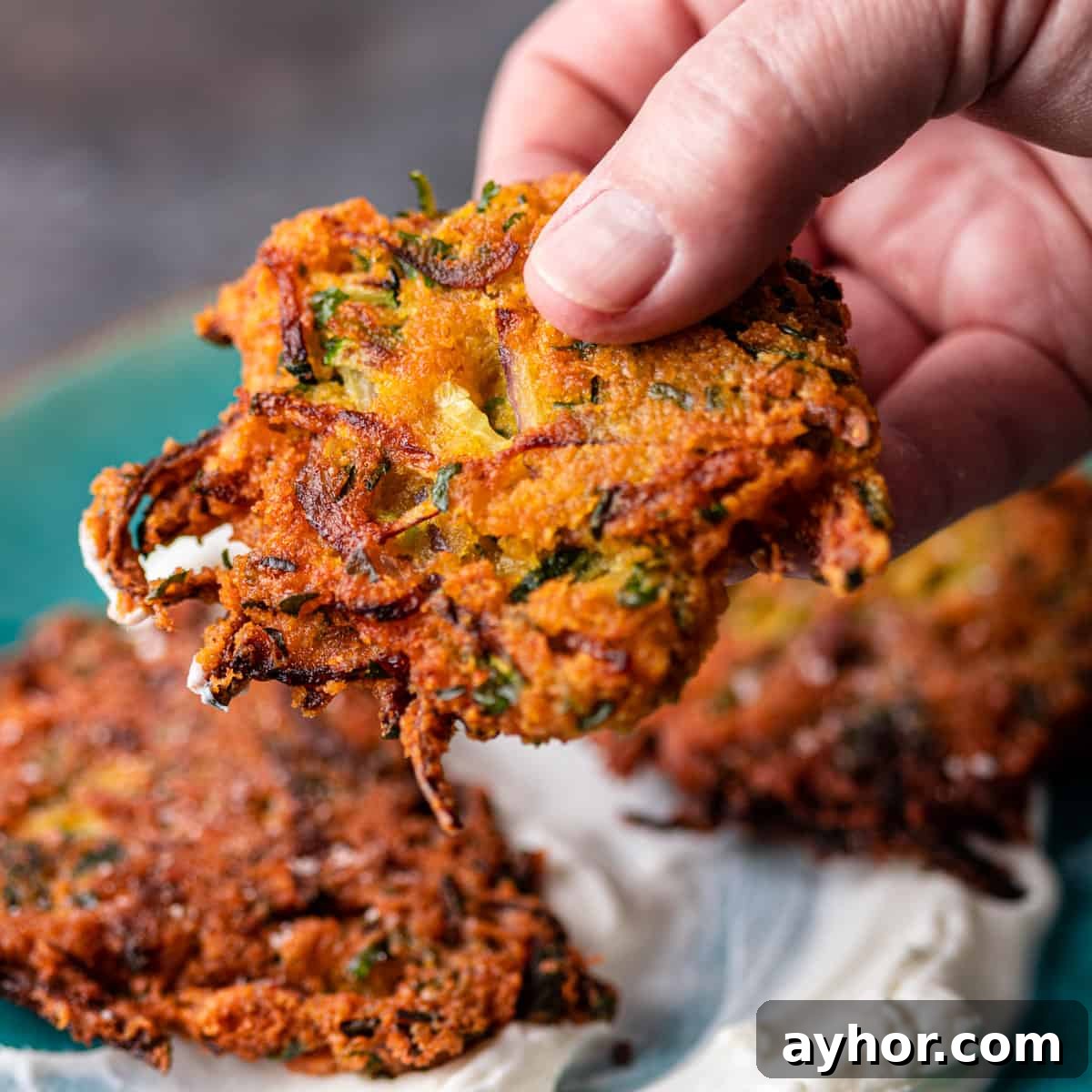 Crispy Zucchini and Lentil Fritters 2 closeup side view: a hand holding zucchini lentil fritters over a plate with yogurt